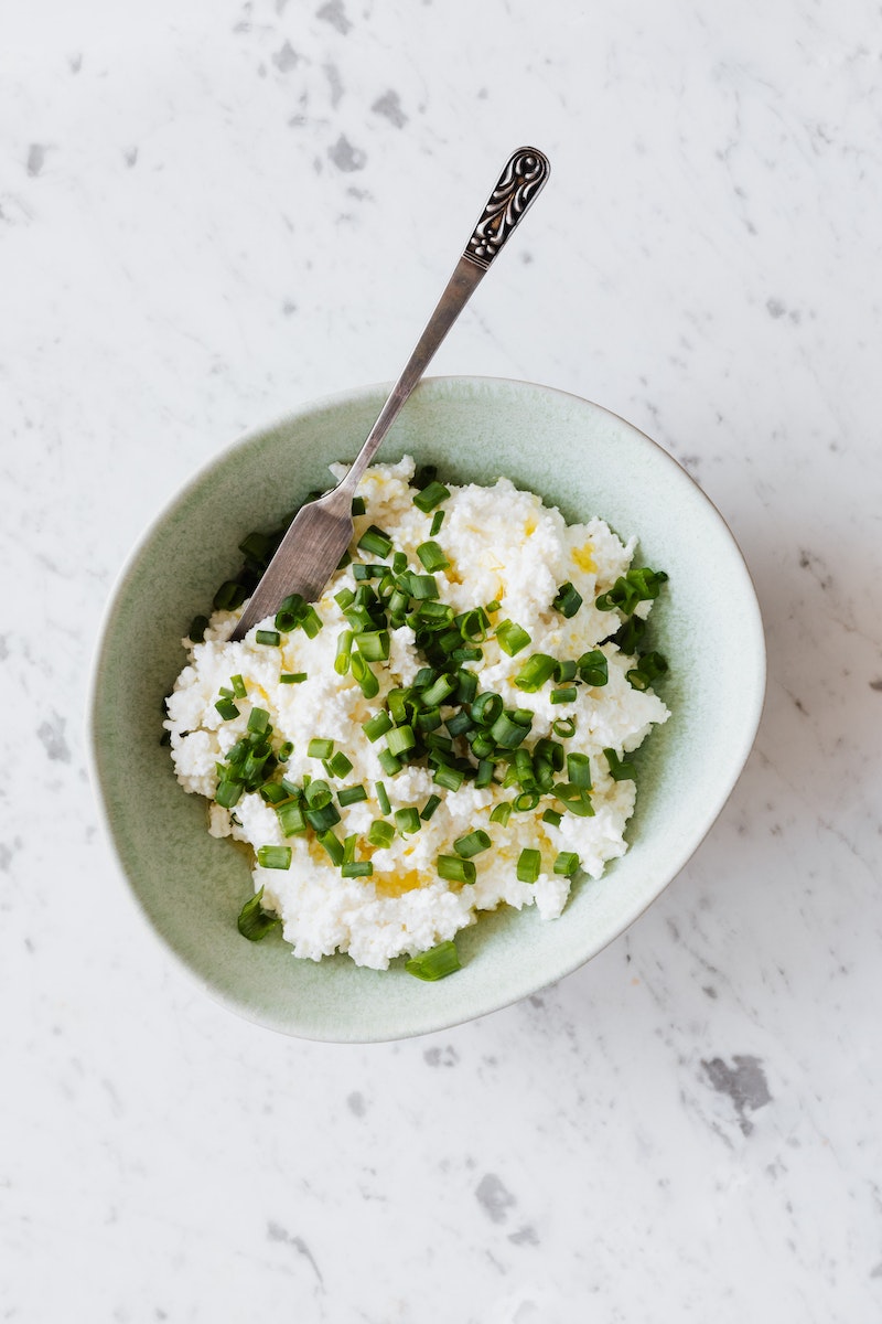 Top view of bowl with cottage cheese with greenery and green onion placed on white table with gray dots and cutlery