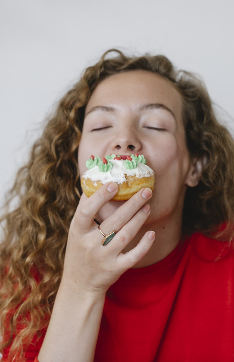Young woman with long curly hair wearing red sweater biting delicious donut with white icing and green cream on white background
